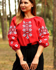Woman wearing a red blouse with white floral embroidery and black pants standing in a forest.