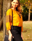Woman in a yellow vyshyvanka and black pants standing next to a tree in an autumn park.