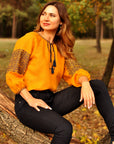 A woman sitting on a tree stump wearing a mustard yellow blouse with dark blue embroidery and black pants.