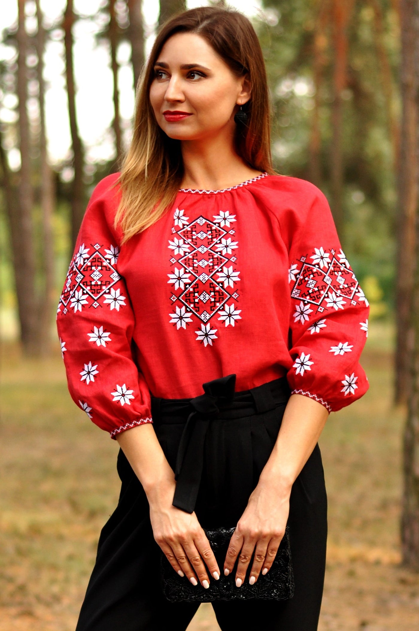 Woman wearing a red blouse with white floral embroidery and black pants standing in a forest.