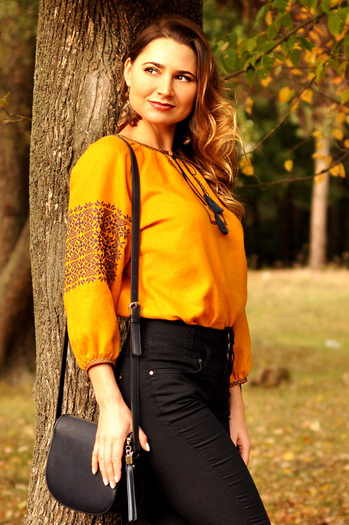 Woman in a yellow vyshyvanka and black pants standing next to a tree in an autumn park.