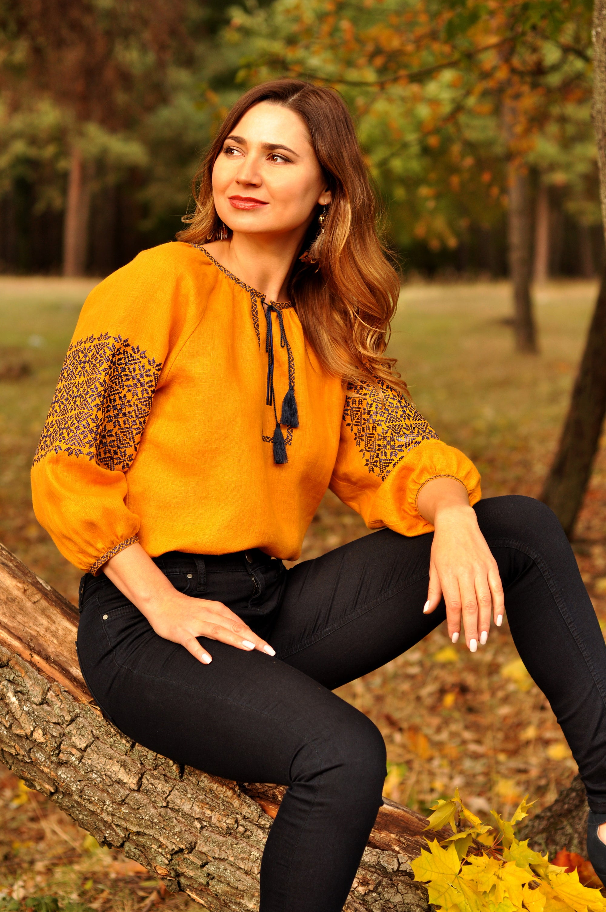 A woman sitting on a tree stump wearing a mustard yellow blouse with dark blue embroidery and black pants.